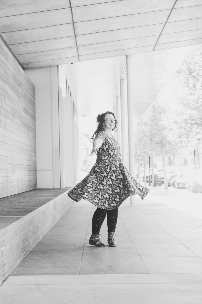 A smiling person wearing a floral dress twirls in an outdoor space with modern architecture.